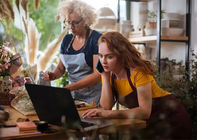 Zwei Floristinnen in einer Blumenwerkstatt – eine arbeitet am Laptop, die andere bindet einen Blumenstrauß.